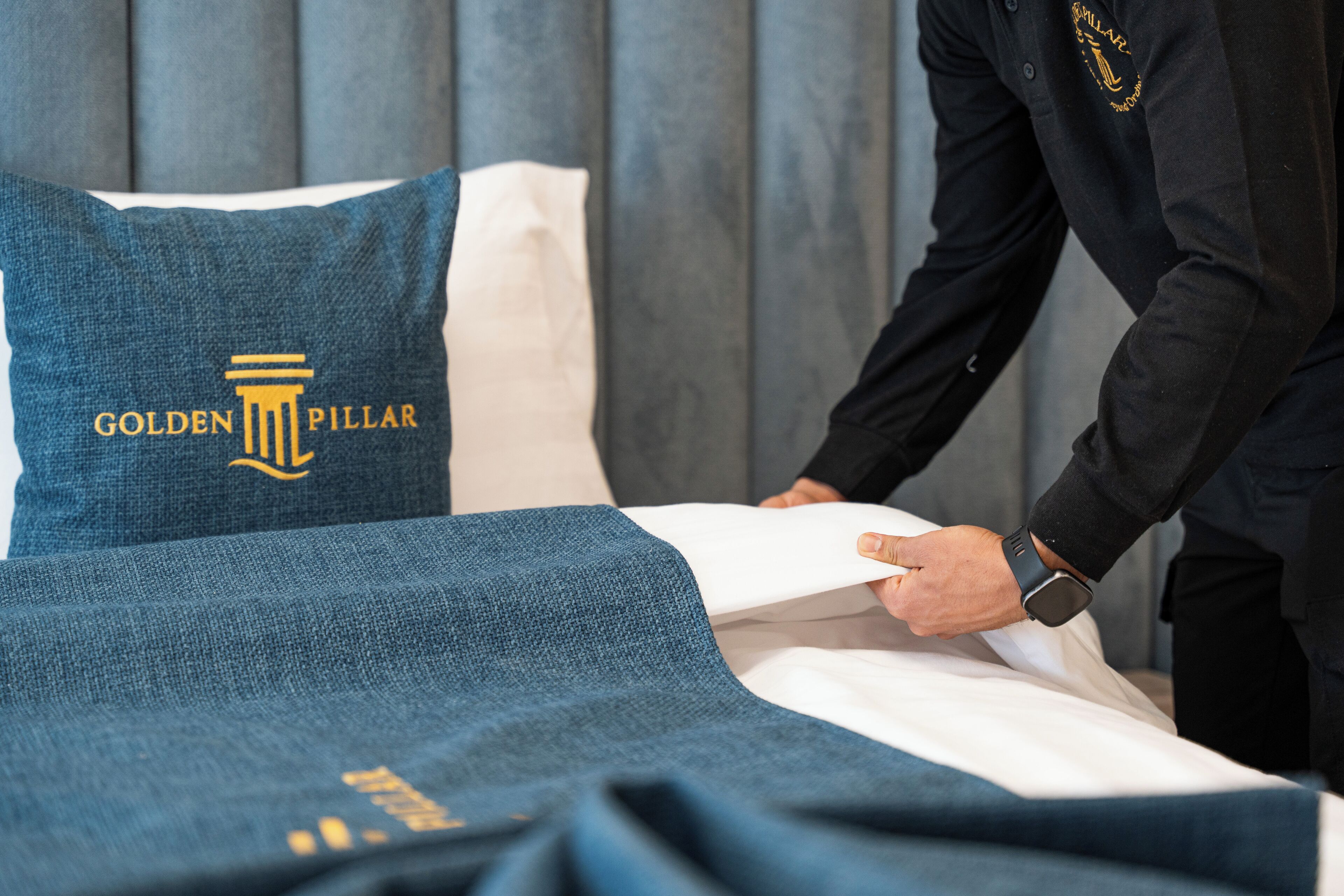 A bed with a pillow featuring the 'Golden Pillar' logo, a person in a suit adjusting the bedspread, and a grey wall in the background.