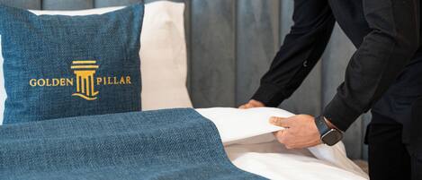 A bed with a pillow featuring the 'Golden Pillar' logo, a person in a suit adjusting the bedspread, and a grey wall in the background.