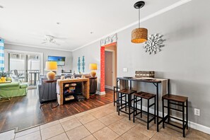 A modern living room with a television, a ceiling fan, and a dining area with stools and a table. - A Place At The Beach II 1F Shore Drive- 2BR- Pool (Myrtle Beach)