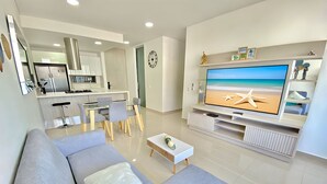 Ein modernes Wohnzimmer mit einem großen Flachbildfernseher, einer Couch mit Zierkissen, einem Couchtisch und einem Essbereich mit Glastisch und Stühlen.