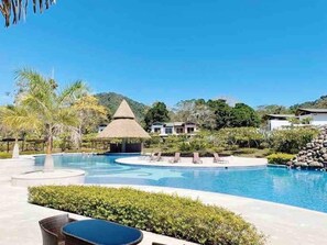 A resort pool area with a thatched-roof structure, lounge chairs, and a clear view of the surrounding landscape. - Gated Tropical Retreat 3BR - Resort Pool, BBQ, walk to beach (Jacó)