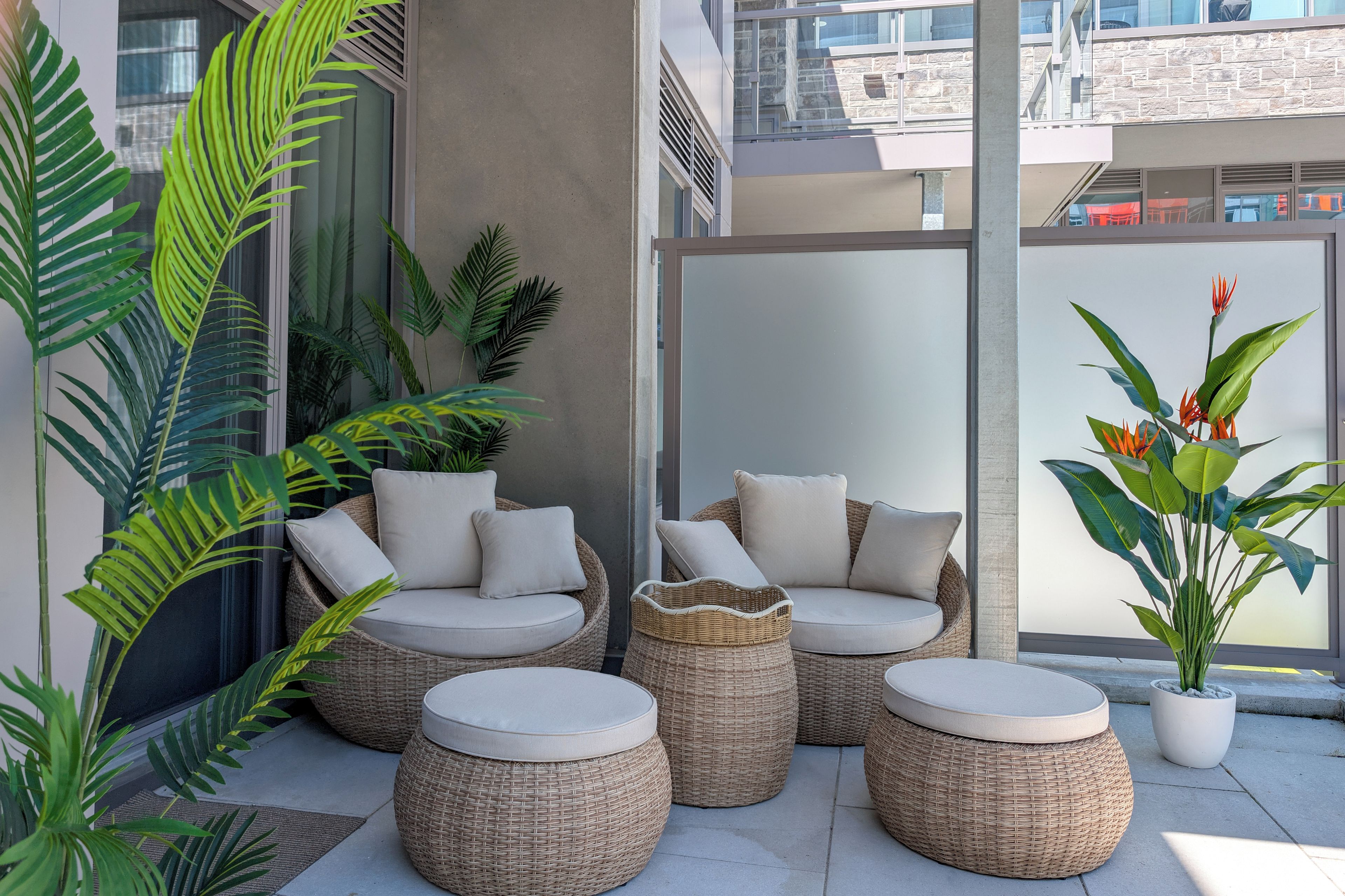 A balcony with wicker furniture, including a sofa and ottomans, surrounded by potted plants.