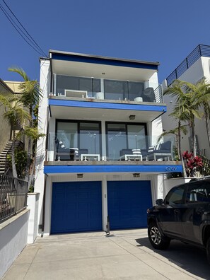 Un edificio de dos plantas con fachada azul y blanca, un balcón con mobiliario exterior y un garaje con dos puertas azules.