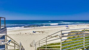 Una playa con una escalera que baja a la orilla, una torre de salvavidas y un cielo azul despejado.