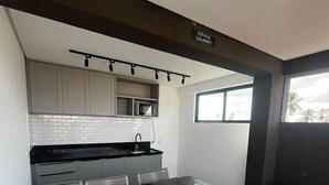 A modern kitchen with a black countertop, white cabinets, and a microwave. There is a dining area with a black table and white chairs. A trash can is visible near the window. - Marina do Cupe 203 (Ipojuca)