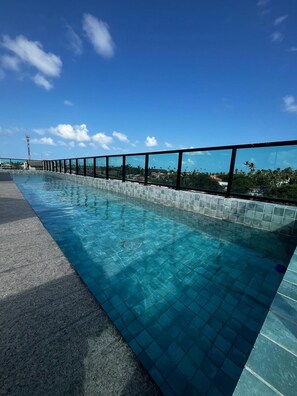 A clear blue swimming pool with tiled edges, surrounded by a glass barrier and a view of palm trees and buildings under a clear sky. - Marina do Cupe 203 (Ipojuca)