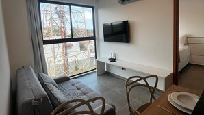A modern living room with a sofa, dining table, chairs, a TV, and a large window overlooking a construction site. - Marina do Cupe 203 (Ipojuca)
