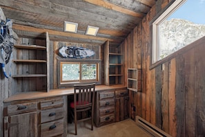 A rustic cabin interior with a wooden desk, a chair, a window with a view of a snowy mountain, and a shelf with a helmet.