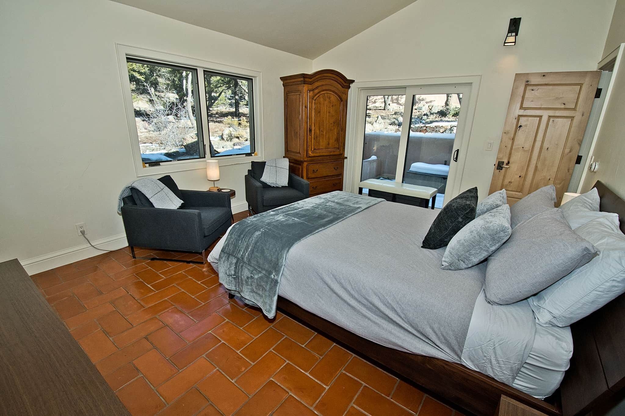 A bedroom with a large bed, two chairs, a wooden wardrobe, and a view of the outdoors through two windows.