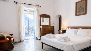 A bedroom with a bed, a dresser, a nightstand, a hanging light, a window with curtains, and a painting on the wall. - Casa Lucia  (Positano)