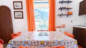 A dining area with a table covered in a patterned tablecloth, orange curtains, and a view of a mountainous landscape. - Casa Lucia  (Positano)