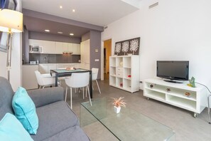Ein modernes Wohnzimmer mit einer Couch, einem Glastisch, einem Fernseher auf einem weißen Schrank und einer Küche im Hintergrund.