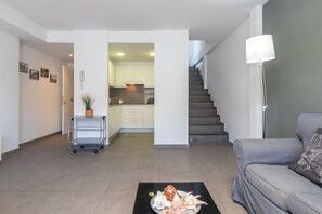 Ein modernes Wohnzimmer mit einem grauen Sofa, einem Couchtisch mit Dekorationsobjekten, einer Treppe und einer Küche im Hintergrund.