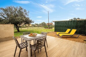 An outdoor patio with a table and chairs, a covered area with yellow lounge chairs, and a view of a green hedge and buildings in the background. - Pals A: Golf en el Jardín y Playa a un Paso (Pals)