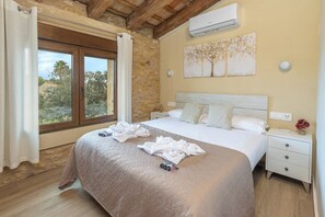 A bedroom with a large bed, stone walls, a wooden ceiling, an air conditioner, a nightstand, and a window with curtains.