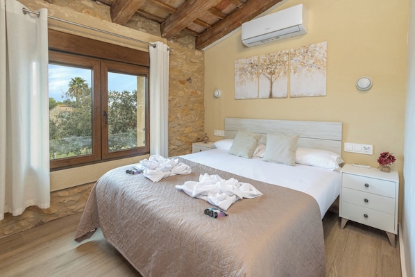 A bedroom with a large bed, stone walls, a wooden ceiling, an air conditioner, a nightstand, and a window with curtains.
