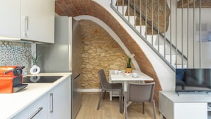 Una cocina compacta con pared de piedra, un comedor con sillas, un televisor y una escalera con barandilla de metal.