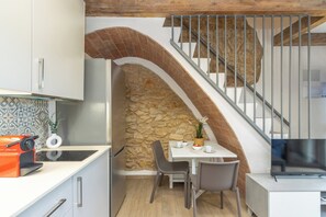 A compact kitchen with a stone wall, a dining area with chairs, a TV, and a staircase with a metal railing.