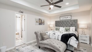 A bedroom with a tufted headboard, a tufted bench, a bed with a patterned bedspread, two bedside lamps, and a ceiling fan. - Gig ‘Em Ags! Pool table, patio, 7 mins Kyle Field (Bryan)