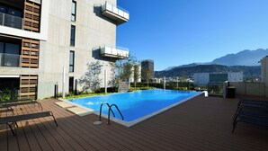 Une piscine sur le toit, dotée d’une terrasse en bois et de rampes en métal, entourée de bâtiments modernes et offrant une vue sur les montagnes.