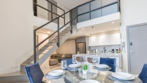 Un espace repas moderne avec une table en verre, des chaises recouvertes d’un tissu bleu et un escalier menant à une mezzanine.
