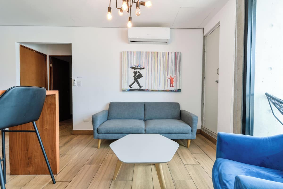 A modern living room with a grey sofa, a white coffee table, and a blue armchair. There is a colorful artwork on the wall and an air conditioning unit above the sofa.