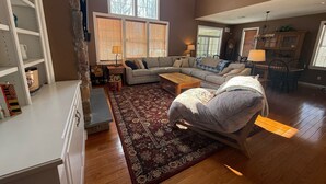 A spacious living room with a large sectional sofa, a coffee table, and a patterned rug. There are multiple windows with blinds, a stone accent wall, and a dining area with a chandelier. - Tremendous 5-bedroom house in Pocono Pines (Pocono Pines)