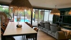 A modern living room with a dining area, a grey sofa, and a pool visible through large windows. - Villa Patio Bleu, heated pool, center & beach (Arès)