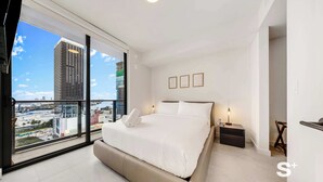 A modern bedroom with a large bed, a nightstand, a lamp, and a view of a cityscape through a large window. - High-Rise Miami Condo | Rooftop Pool | Sea View (Miami)