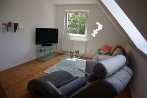 Een moderne woonkamer met een flatscreen televisie, een glazen salontafel, een bank en een raam met uitzicht op groen.