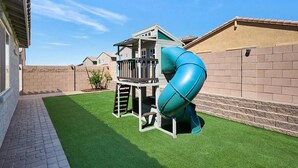 Un'area giochi con scivolo e struttura per arrampicata in un giardino privato con prato sintetico e muretto in mattoni.