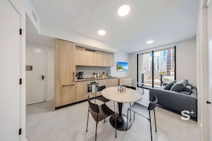 A modern kitchen with wooden cabinets, a dining table with chairs, and a sofa in a living area with a large window. - Rooftop Pool Oasis in Downtown Miami Condo w/ Gym (Miami)