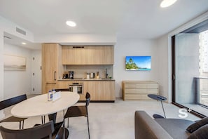 A modern kitchen with wooden cabinets, a dining table with chairs, a sofa, and a view of a building through a large window. - Rooftop Pool Oasis in Downtown Miami Condo w/ Gym (Miami)