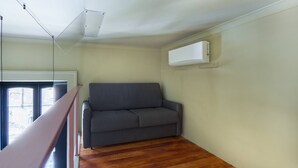 Een kamer met een grijze bank, houten vloer, een wit plafond en een raam met blinds.