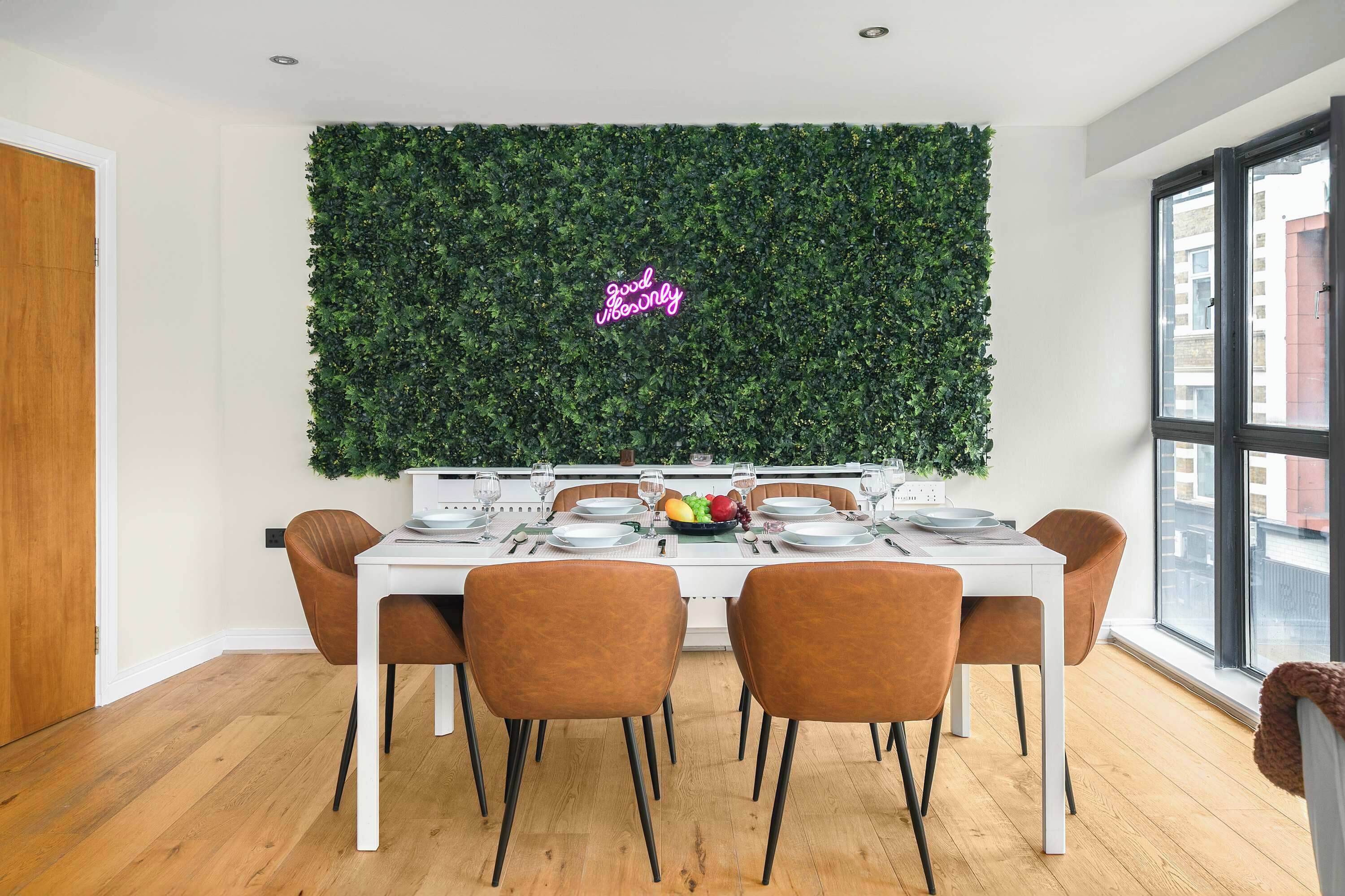 Een eethoek met een witte tafel gedekt voor vier personen, omringd door bruine stoelen, een groene muur met een neonbord, en een raam met uitzicht op gebouwen.