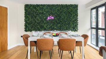 Een eethoek met een witte tafel gedekt voor vier personen, omringd door bruine stoelen, een groene muur met een neonbord, en een raam met uitzicht op gebouwen.