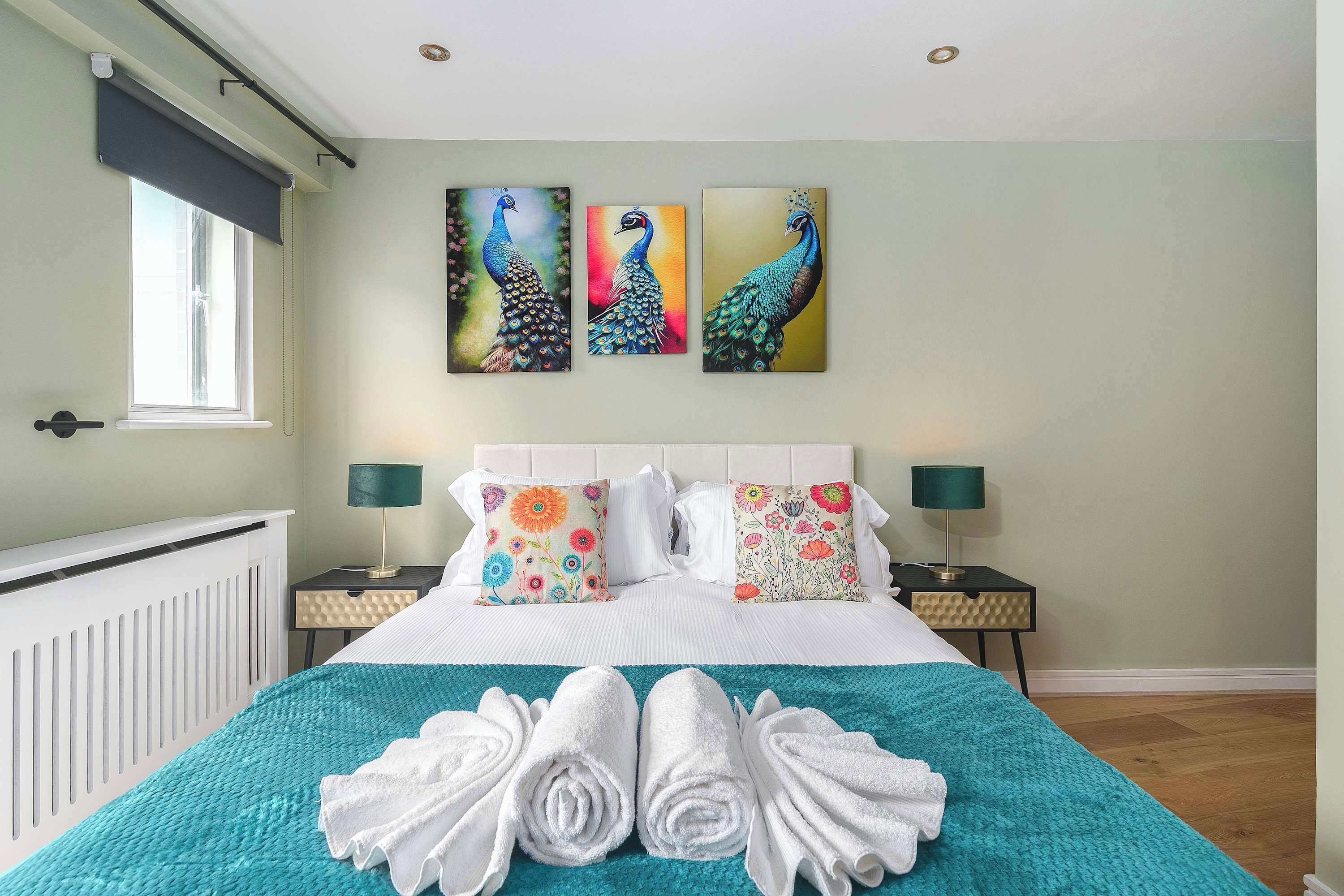 A hotel room with a bed, two nightstands, a window, and three peacock paintings on the wall.