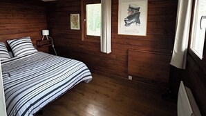 A wooden-paneled bedroom with a bed, a nightstand, a framed picture, and a window with curtains. - Chalet in the woods (Meyreuil)