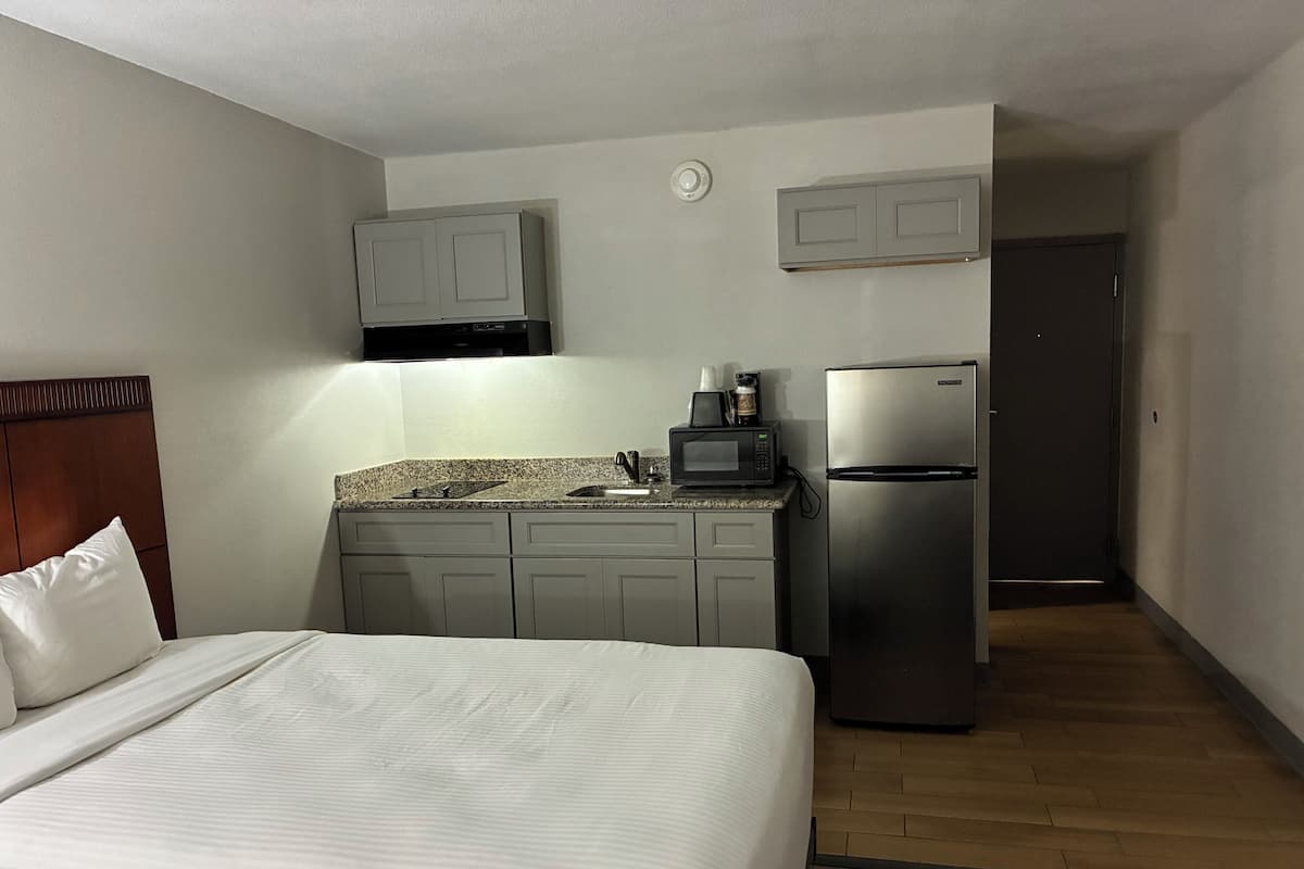 A hotel room with a bed, a kitchenette with a microwave, a refrigerator, and a door leading to another room.