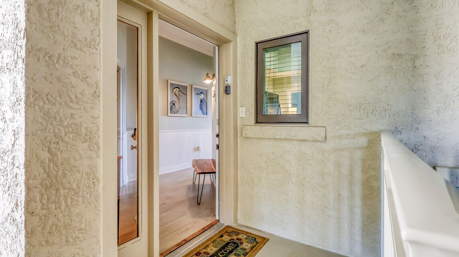 Un pasillo con piso de madera, una puerta que da a otra habitación y una pequeña mesa con una silla.