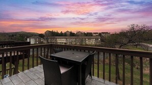 Un balcone con tavolo e sedie, affacciato su una zona residenziale con case e alberi al tramonto.