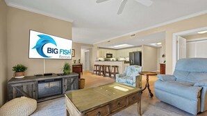 Ein Wohnzimmer mit Sofa, Sessel und Couchtisch. Im Hintergrund ist eine Küche mit Mikrowelle und Kühlschrank zu sehen.