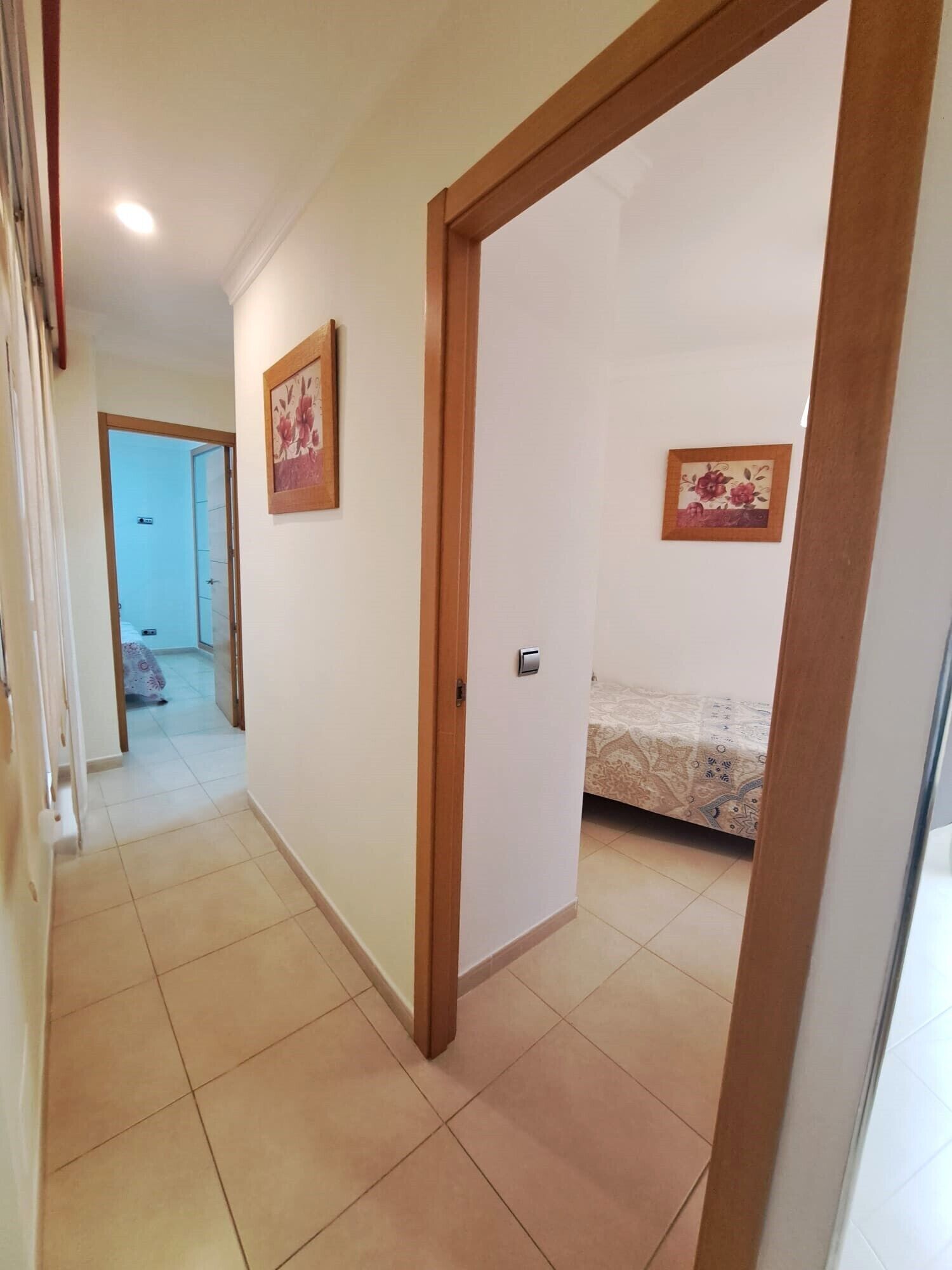 A hallway with a bed in the background, a door frame, and two framed pictures on the wall.