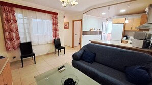 Una sala de estar con un sofá, dos sillones, una mesa de centro con un cuenco, y una zona de cocina con refrigerador, microondas y estufa.