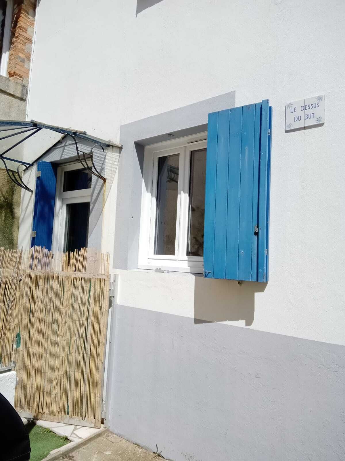 A white building with a small window and a blue shutter. There is a sign on the wall that reads "LE DESSUS DU BOUT."