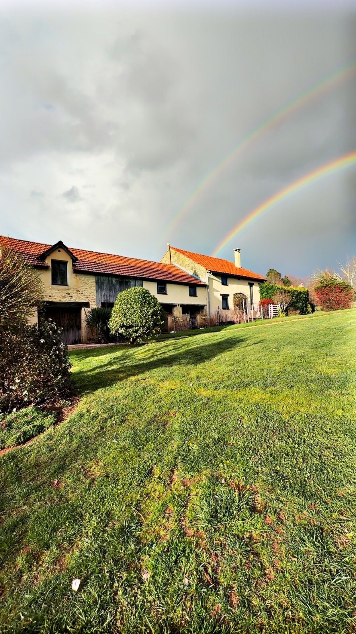 Een huis met een rood dak, een grote voortuin en een regenboog aan de lucht.