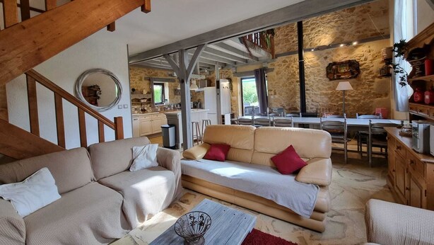 Amplia sala de estar con techo alto, vigas a la vista y pared de piedra. Una escalera de madera conduce a un nivel superior. La habitación cuenta con un sofá grande, una mesa de centro y un comedor con mesa y sillas.