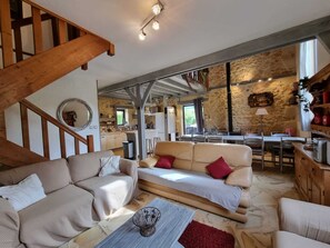A spacious living area with a high ceiling, exposed beams, and a stone wall. A wooden staircase leads to an upper level. The room features a large sofa, a coffee table, and a dining area with a table and chairs. - Near Sarlat and Rocamadour, large vacation rental, sleeps 10 with a private pool (Rouffilhac)