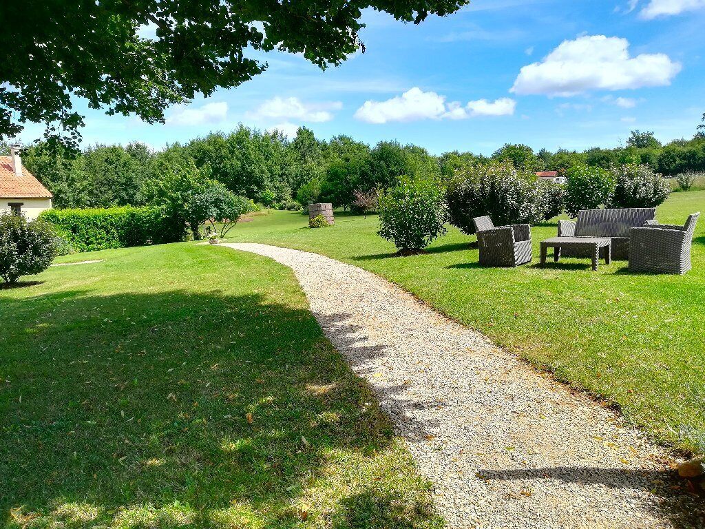 Un sendero de grava conduce a muebles de mimbre en un jardín exuberante y verde, con una casa al fondo.