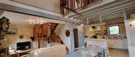 A spacious living area with a high ceiling, a wooden staircase, a dining table, and a kitchen area.